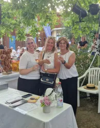 Drei Frauen posieren lächelnd an einem Tisch, während sie ein Getränk halten. Im Hintergrund sind Gäste und eine gemütliche Outdoor-Atmosphäre zu sehen.