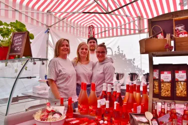 Ein lebhaftes Marktstand-Team steht unter einem gestreiften Zelt. Im Vordergrund sind Flaschen mit rotem Getränk und Snacks sichtbar.