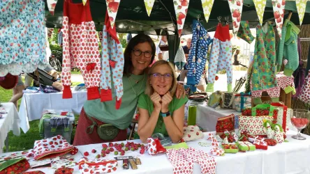 Zwei lächelnde Frauen stehen hinter einem Verkaufsstand mit bunten Stoffen und kleinen Accessoires. Der Stand ist mit fröhlichen Stoffen in verschiedenen Farben dekoriert.