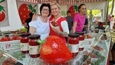 Ein lebendiger Erdbeerstand mit frischen Erdbeeren und zwei lächelnden Verkäuferinnen. Im Vordergrund steht eine große, dekorative Erdbeere und mehrere Gläser mit Erdbeermarmelade.