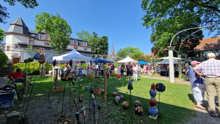 Ein lebhafter Markt im Freien mit bunten Ständen und vielen Besuchern. Umgeben von Bäumen und einer schönen Architektur bietet der Platz eine freundliche Atmosphäre.