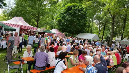 Eine belebte Veranstaltung im Freien mit vielen Menschen. Es gibt verschiedene Stände und Tische unter Bäumen.