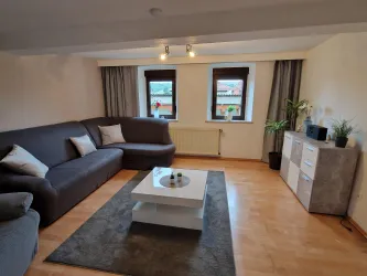 Ein modernes Wohnzimmer mit einem grauen Sofa, einem weißen Couchtisch und einer gemütlichen Atmosphäre. Große Fenster lassen viel Tageslicht herein.