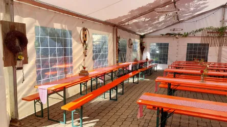 Ein Festzelt mit langen, orangefarbenen Bänken und Tischen. Die Wände sind mit großen Fenstern und einfachen Dekorationen geschmückt.