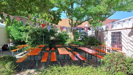 Ein gemütlicher Außenbereich mit länglichen Tischen und Bänken, umgeben von grünen Pflanzen. Bunte Fahnen hängen über dem Platz und schaffen eine festliche Atmosphäre.