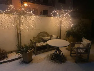Ein schöner Wintergarten mit einer gemütlichen Sitzgruppe und einem runden Tisch. Die Bäume sind mit Lichtern dekoriert und der Boden ist mit Schnee bedeckt.