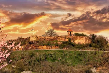 Eine alte Ruine auf einem Hügel, umgeben von Bäumen und Blumen. Der Himmel ist dramatisch mit orangefarbenen und violetten Wolken.