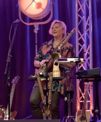 Eine Musikerin steht auf einer Bühne und spielt Saxophon. Sie trägt ein buntes Hemd und hat eine fröhliche Ausstrahlung.