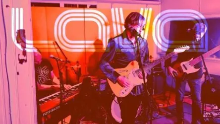 Eine Band spielt live in einem kreativen Raum. Die elektrische Atmosphäre wird durch die farbenfrohe Beleuchtung und das Wort "LOVE" verstärkt.