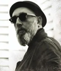 Ein Mann mit grauem Bart, Sonnenbrille und schwarzem Hut schaut zur Seite. Der Hintergrund ist unscharf und hell.