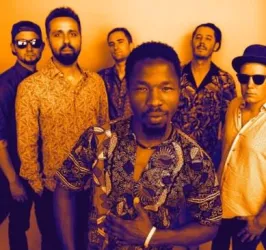 Eine Gruppe von sechs Männern posiert vor einem orangefarbenen Hintergrund. Sie tragen farbenfrohe Hemden und zeigen eine freundliche, entspannte Haltung.