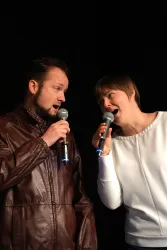Ein Mann und eine Frau stehen auf der Bühne und singen miteinander in ein Mikrofon. Beide haben Spaß und sind konzentriert auf ihre Darbietung.
