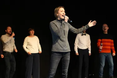 Eine Gruppe von fünf Personen steht auf einer Bühne und singt. Eine Frau mit einem Mikrofon führt den Gesang an, während die anderen sie unterstützen.