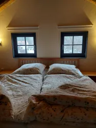 Ein gemütliches Schlafzimmer mit einem großen Bett und hellen Bettdecken. Zwei Fenster bieten einen Blick nach draußen.