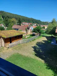 Eine idyllische Landschaft mit einem kleinen Dorf und grünen Wiesen. Im Vordergrund steht eine Scheune und Bäume schützen die Umgebung.