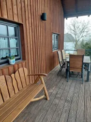 Eine gemütliche Terrasse aus Holz mit einem Holztisch und Stühlen. Im Hintergrund sind eine Wand aus Holz und einige Fenster zu sehen.