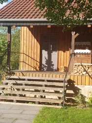 Ein Holzhaus mit einer Treppe und einer grauen Tür. Der Garten ist gepflegt und im Hintergrund sind Holzscheite gestapelt.