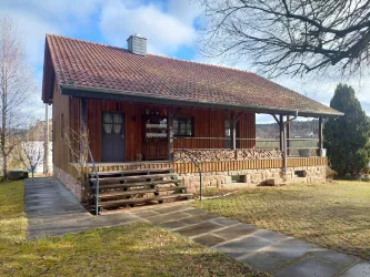 Ein hübsches Holzhaus mit einer Veranda und einem roten Dach. Die Umgebung ist grün und einladend, ideal für entspannte Tage im Freien.