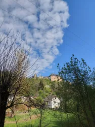 Eine Burg thront auf einem Hügel unter einem blauen Himmel mit Wolken. Im Vordergrund sind Bäume und ein kleines haus zu sehen.