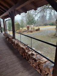 Ein überdachter Holzsteg mit einem Sichtschutz aus aufgeschichtetem Holz. Im Hintergrund sind Bäume und ein gepflegter Garten zu sehen.