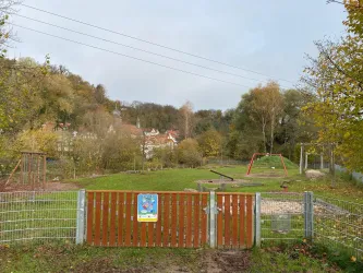 Ein Spielplatz mit einer grünen Wiese und einigen Spielgeräten. Der Bereich ist mit einem Zaun und einem Tor umgeben, umgeben von Bäumen und Hügeln.
