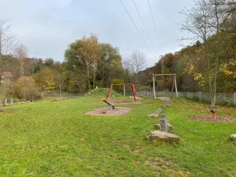 Ein Spielplatz mit einer Schaukel und einer Wippe in einer grünen Umgebung. Bäume und eine friedliche Landschaft umgeben den Spielbereich.