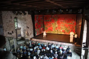 Eine Theateraufführung in einem historischen Gebäude. Auf der Bühne stehen zwei Darsteller vor einem bunten Hintergrund, während das Publikum zuschaut.