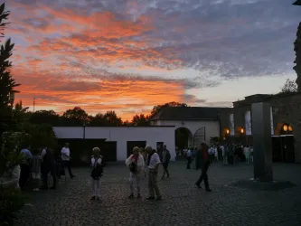 Eine Gruppe von Menschen steht auf einem Platz bei Sonnenuntergang. Der Himmel ist in warmen Farben gefärbt und schafft eine einladende Atmosphäre.
