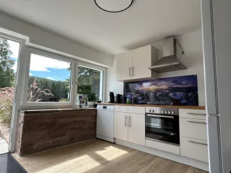 Eine moderne Küche mit weißen Schränken und Holzoberflächen. Große Fenster bringen viel Tageslicht und bieten einen Blick nach draußen.