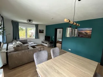 Ein modernes Wohnzimmer mit einem bequemen Sofa und einem Esstisch. Die Wände sind in einem beruhigenden Türkis und es gibt große Fenster, die viel Licht hereinlassen.