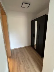 Ein kleiner Raum mit einem schwarzen Kleiderschrank und einem hellen Holzboden. Die Wände sind weiß und es gibt eine Deckenleuchte.