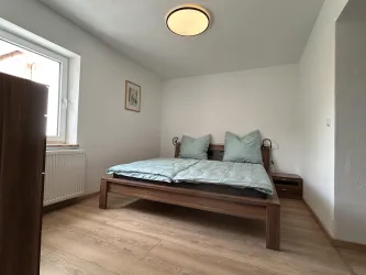 Ein einfaches Schlafzimmer mit einem Bett und zwei Kissen in hellblauer Farbe. Der Raum hat einen hellen, freundlichen Look durch das Fenster und den Holzfußboden.