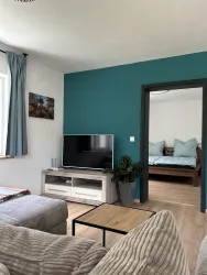 Ein gemütlicher Wohnraum mit einem modernen Fernseher und einem bequemen Sofa. Im Hintergrund ist ein Schlafzimmer mit einem Holzbett und grünen Kissen sichtbar.