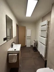 Ein modernes Badezimmer mit einem weißen Waschbecken und einem großen Spiegel. An der Wand befindet sich ein Handtuchheizkörper und der Boden ist mit braunen Fliesen ausgelegt.