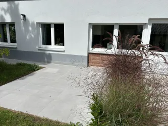 Ein gepflegter Außenbereich mit einem gepflasterten Gehweg und grünen Pflanzen. Die Wand des Hauses ist schlicht und modern.