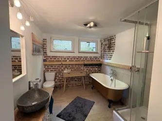 Ein modernes Badezimmer mit einer freistehenden Badewanne und einer separaten Dusche. Die Wände sind mit roten Ziegelsteinen verkleidet, und es gibt viel Tageslicht durch die Fenster.