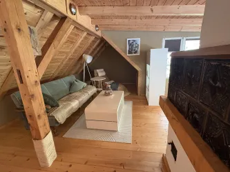 Ein gemütliches Wohnzimmer im Dachgeschoss mit Holzbalken und einem großen Fenster. Das Zimmer ist mit einem Sofa, einem Tisch und einer Lampe ausgestattet.