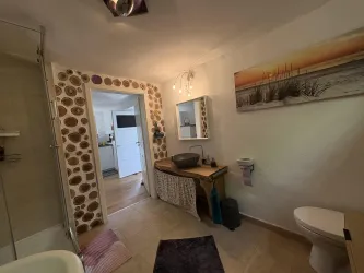 Ein modern gestaltetes Badezimmer mit einer Dusche, einem Waschbecken auf einem Holztisch und einer Toilette. An der Wand hängt ein buntes Bild mit einem Meer und Strandmotiv.