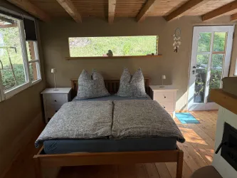 Ein gemütliches Schlafzimmer mit einem großen Bett und zwei Kissen. Große Fenster lassen viel Licht und Blick auf die grüne Umgebung herein.