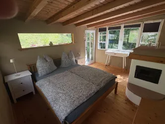 Ein gemütliches Schlafzimmer mit einem großen Bett und Holzdecken. Große Fenster sorgen für viel Tageslicht und Blick ins Grüne.