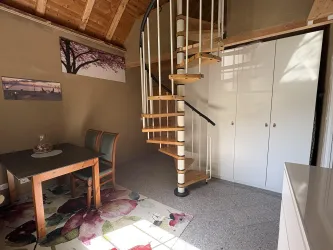 Ein heller Raum mit einer Holztreppe, einem Tisch und Stühlen. An der Wand hängen Bilder und ein großer Kleiderschrank ist zu sehen.