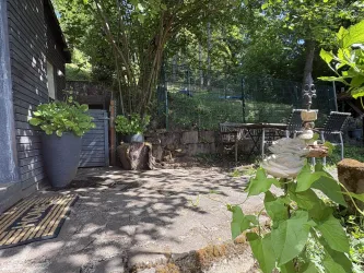 Ein gemütlicher Außenbereich mit einer Terrasse und grünen Pflanzen. Im Hintergrund steht ein Tisch mit Stühlen und es gibt Schatten von Bäumen.