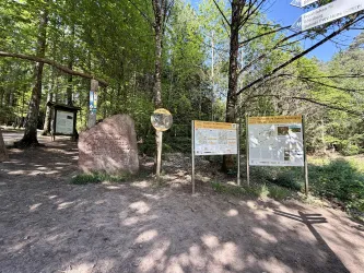 Ein Wanderweg-Eingang im Wald mit mehreren Schildern und Informationen. Im Hintergrund sind grüne Bäume und blauer Himmel zu sehen.