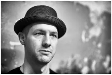 Ein Mann mit einem schwarzen Hut blickt nachdenklich in die Kamera. Der Hintergrund ist unscharf und scheint künstlerisch gestaltet zu sein.