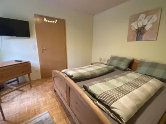 Ein gemütliches Schlafzimmer mit einem Doppelbett und karierten Bettdecken. An der Wand hängt ein Gemälde von weißen Blumen, und es gibt einen Fernseher sowie einen Schreibtisch im Raum.