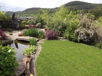 Ein schöner Garten mit grünem Rasen, bunten Pflanzen und einem kleinen Teich. Im Hintergrund sind Hügel und Bäume zu sehen.