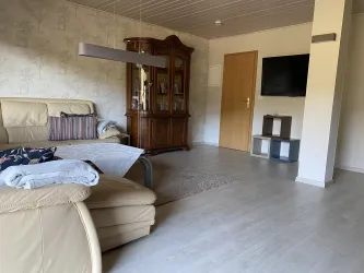 Ein gemütliches Wohnzimmer mit einem beigen Sofa und einem Holzschrank. An der Wand hängt ein Fernseher, und der Boden ist mit einem hellen Teppich bedeckt.