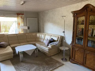 Ein gemütliches Wohnzimmer mit einer beigen, großen Sofaecke und einem Holzhängeschrank. Ein Tisch mit einer weißen Tischdecke steht in der Mitte auf einem beigen Teppich.