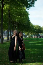 Zwei Musikerinnen stehen auf einer grünen Wiese unter Bäumen. Eine von ihnen hält eine Violine und beide tragen elegante, schwarze Kleider.
