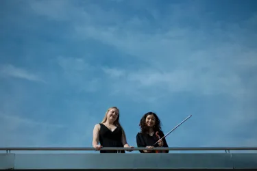 Zwei Frauen stehen auf einem Balkon und lächeln in die Kamera. Der Himmel ist klar und blau.